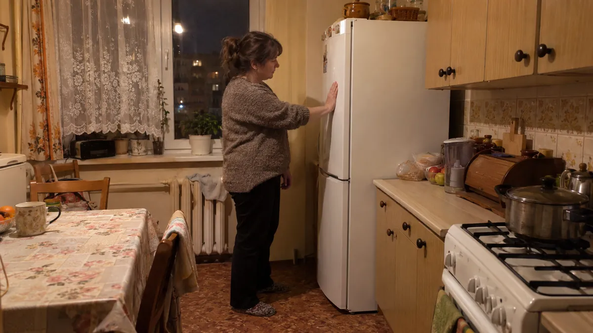 Kobieta w kuchni w bloku wieczorem przykłada dłoń do boku białej lodówki